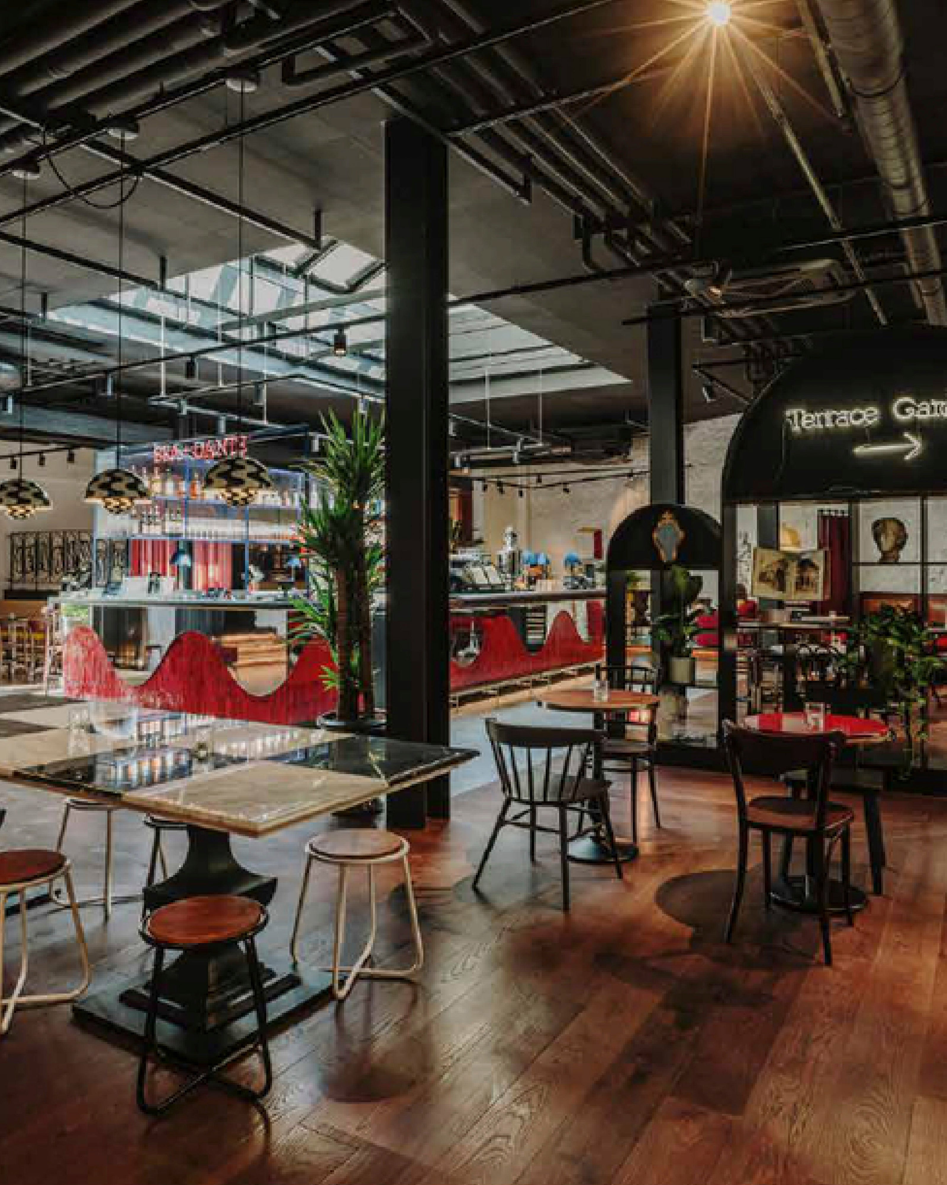 Interior de un restaurante de estilo industrial con techos altos y tuberías expuestas, que muestra mesas de madera, una barra con detalles rojos al fondo y un sistema de iluminación y audio profesional integrado en la estructura del techo.