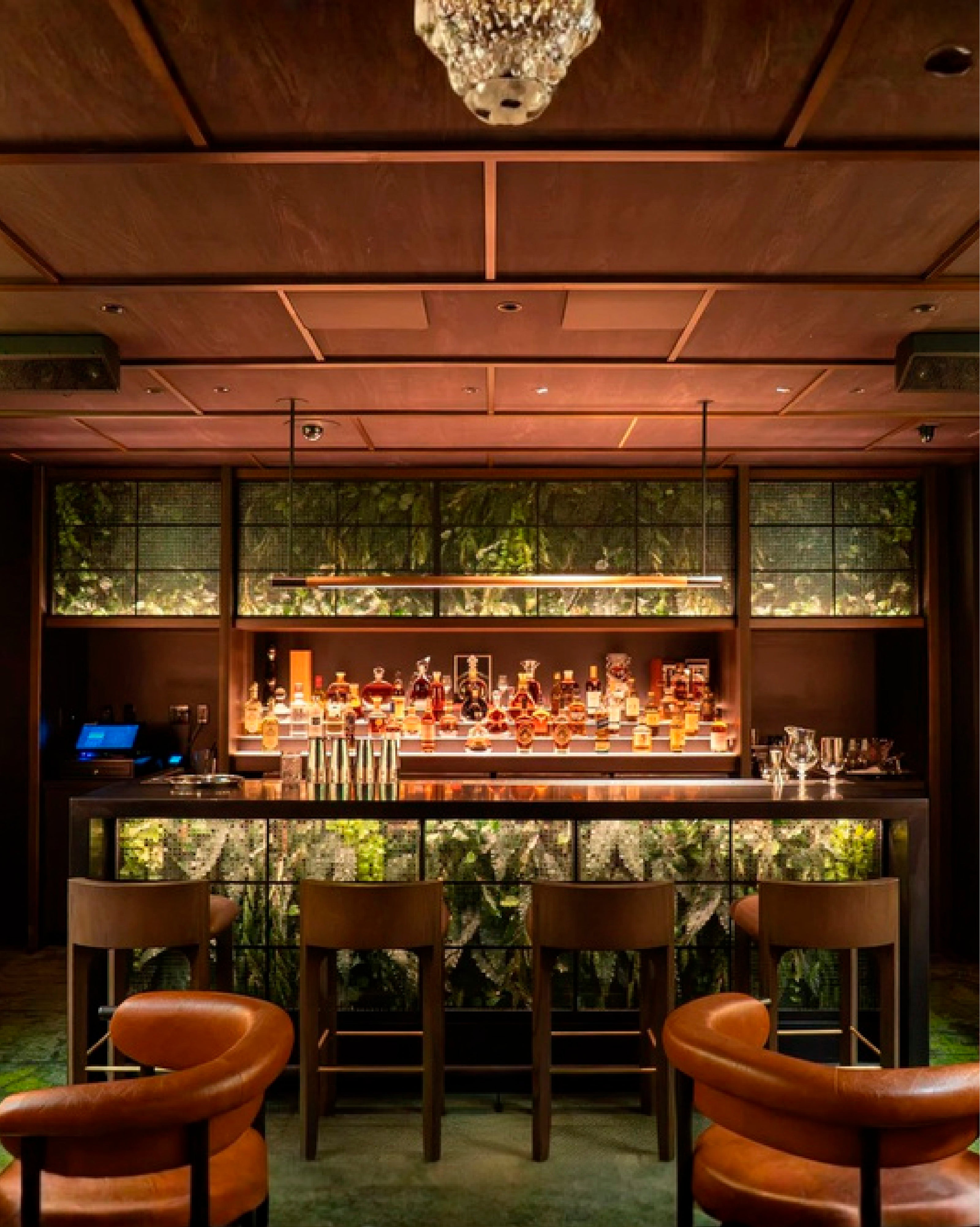Bar de lujo con iluminación tenue y acabados en madera, que muestra una barra con exhibición de botellas iluminadas, sillas de piel color canela y altavoces profesionales de color oscuro montados discretamente en el techo artesonado.