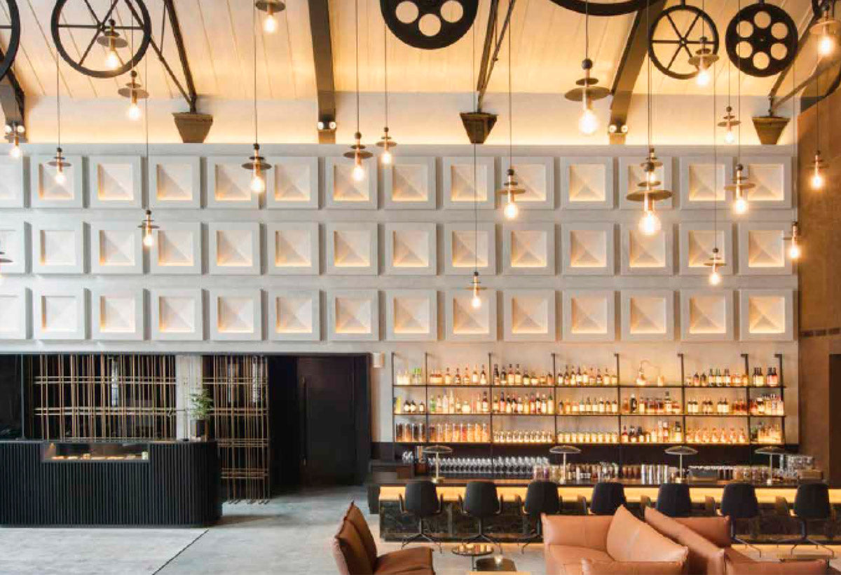 Lobby bar de diseño industrial con una pared de paneles geométricos retroiluminados, que muestra lámparas colgantes tipo Edison, una barra de bar con estantería abierta e iluminada, y altavoces profesionales de pared integrados discretamente bajo las vigas del techo.