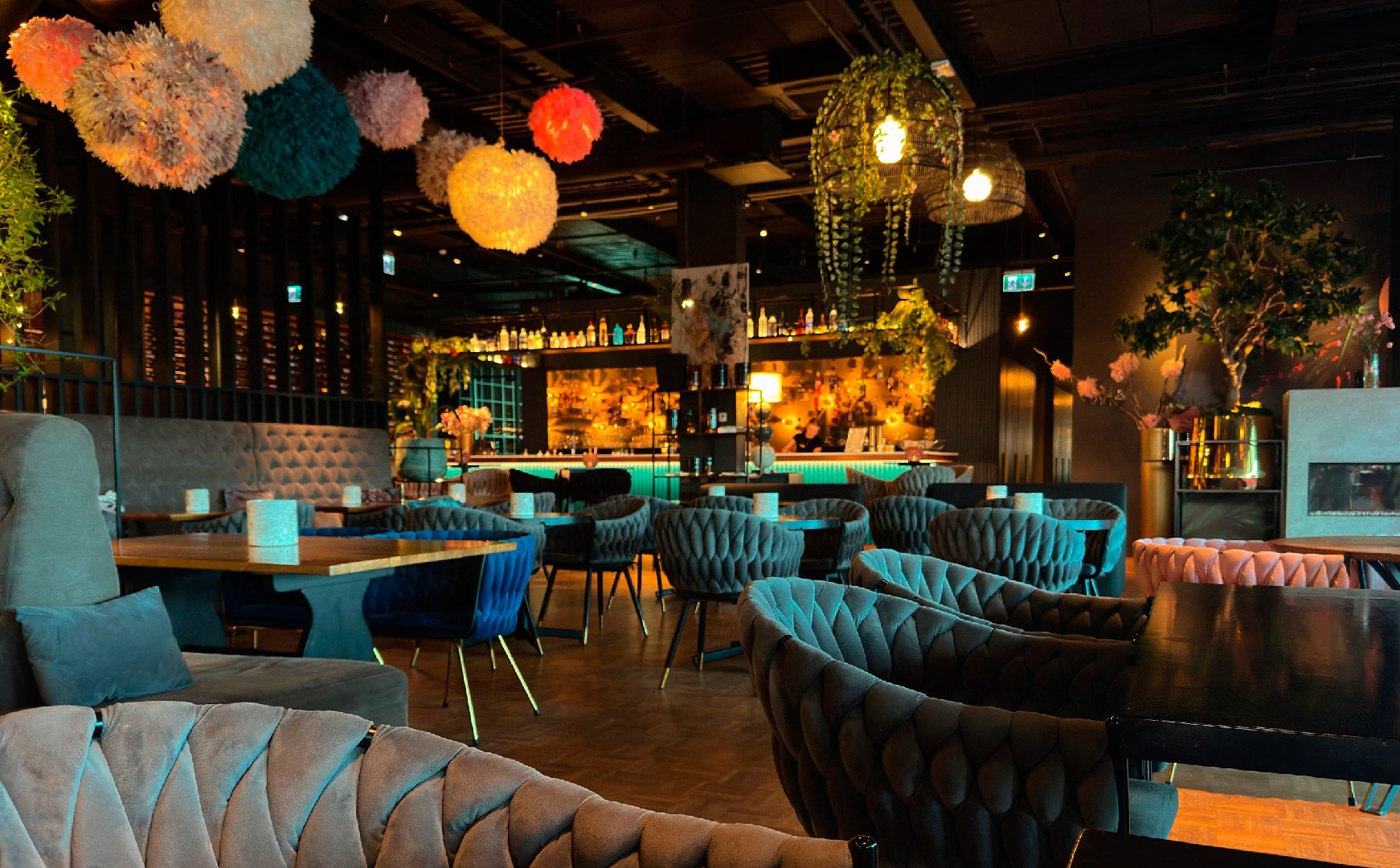 Interior de un restaurante moderno y elegante con mobiliario aterciopelado en tonos grises y azules, decorado con lámparas colgantes esféricas de colores y plantas, mostrando al fondo una barra de bar iluminada en tonos cálidos.