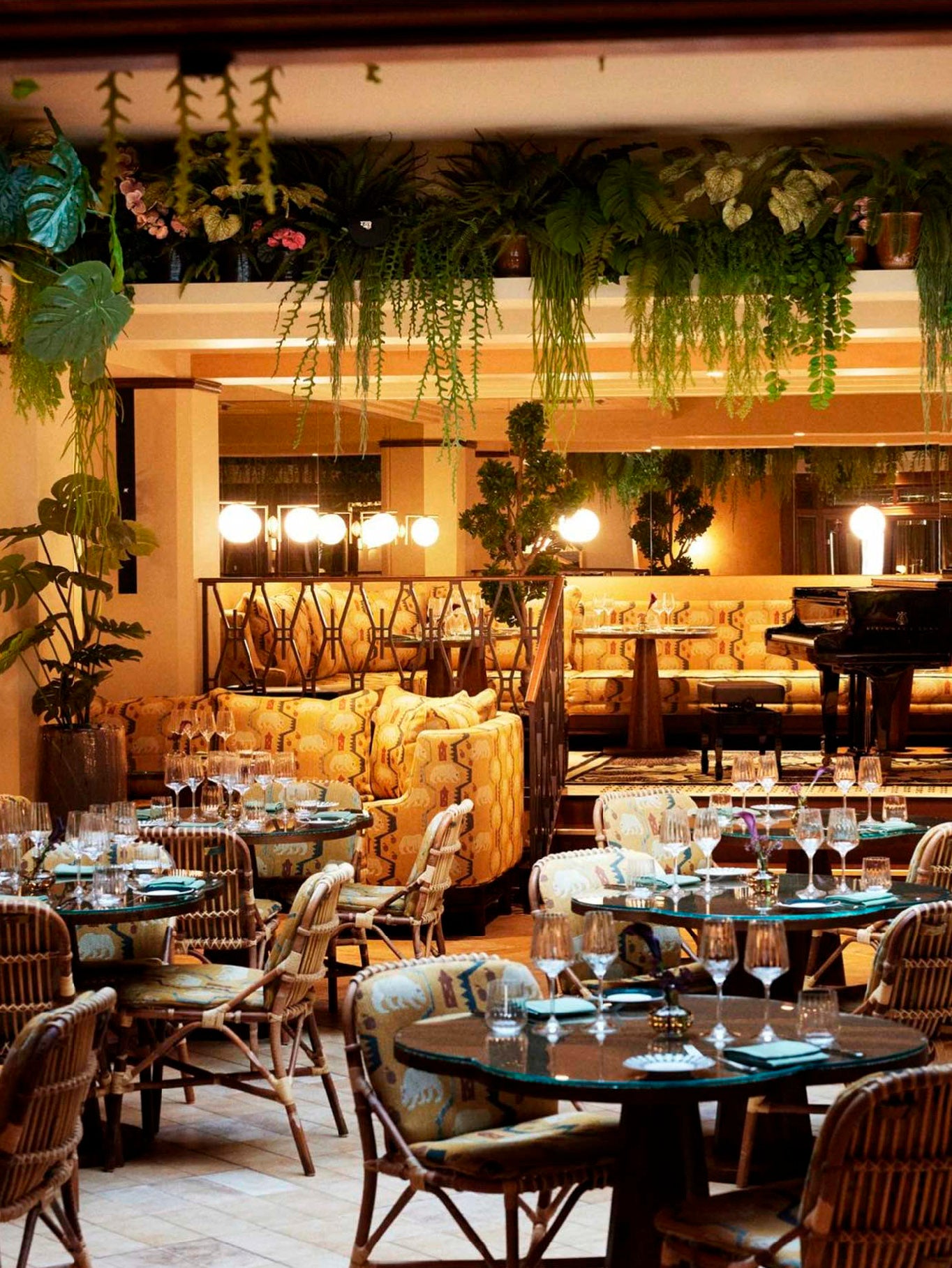 Interior de un restaurante con decoración biofílica, que muestra mesas de cristal, sillas de mimbre, abundante vegetación colgante del techo y un piano de cola al fondo, creando una atmósfera elegante para sistemas de audio ambiental.