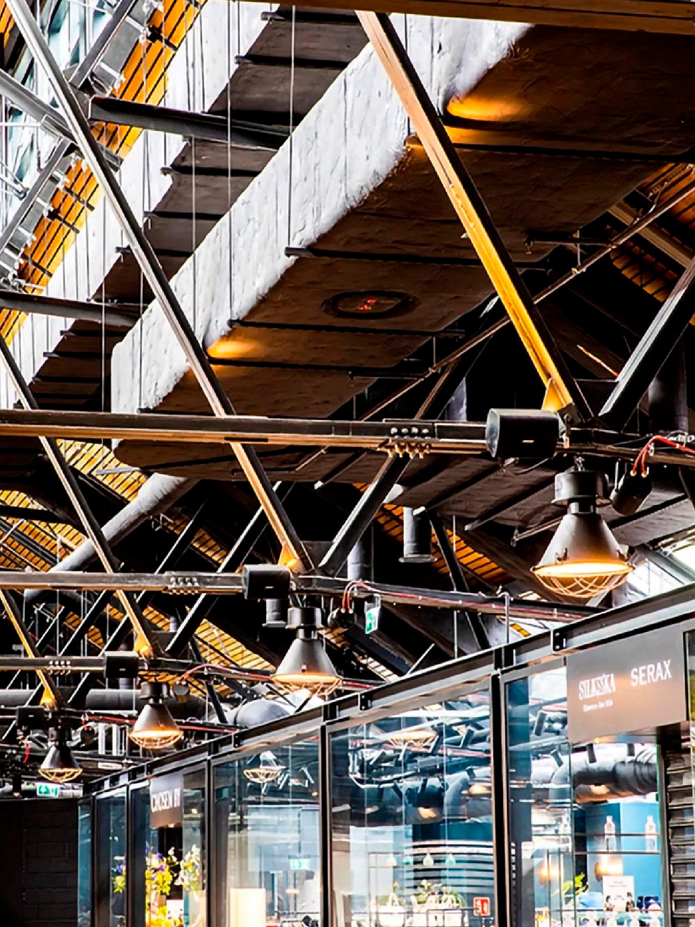 Techo industrial de un centro comercial o tienda retail con vigas de acero expuestas, que muestra lámparas colgantes de estilo rústico y pequeños altavoces profesionales negros instalados estratégicamente para el sistema de audio ambiental.