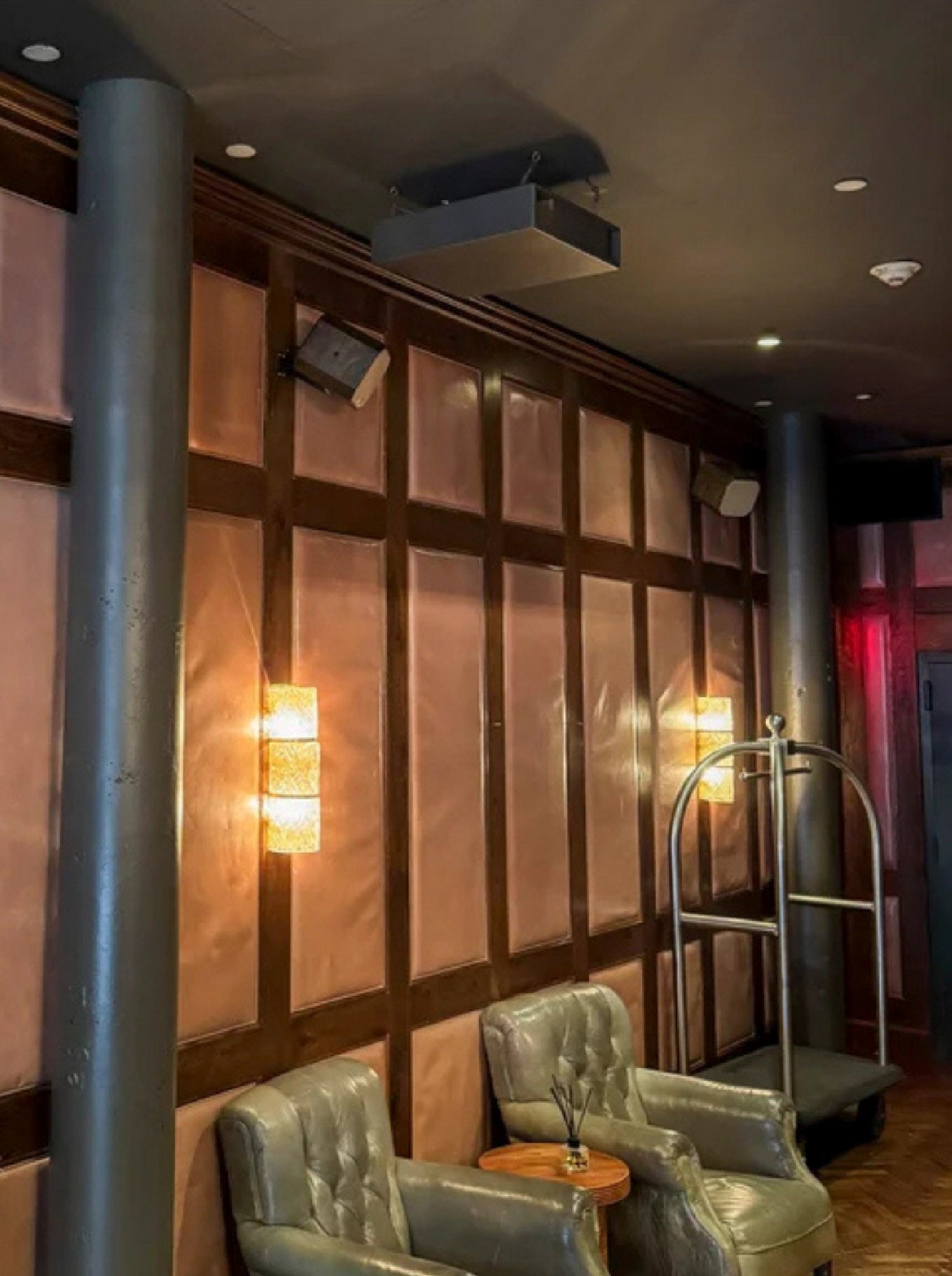 Lobby de hotel con paredes de paneles de madera y piel, que muestra sillones clásicos, un carrito de equipaje y altavoces profesionales de formato pequeño montados en la parte superior de la pared para audio ambiental.